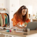 Logistics, laptop and fashion manager of retail or boutique shop checking clothing stock in boxes on a checklist. Small business, entrepreneur and young woman working with clothes inventory in store