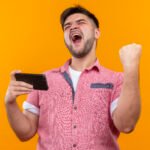 young handsome guy wearing pink polo shirt rejoicing in victory holding phone making did it sign with raised fist standing over orange background
