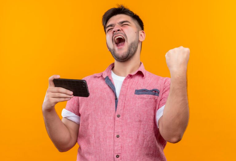 young handsome guy wearing pink polo shirt rejoicing in victory holding phone making did it sign with raised fist standing over orange background